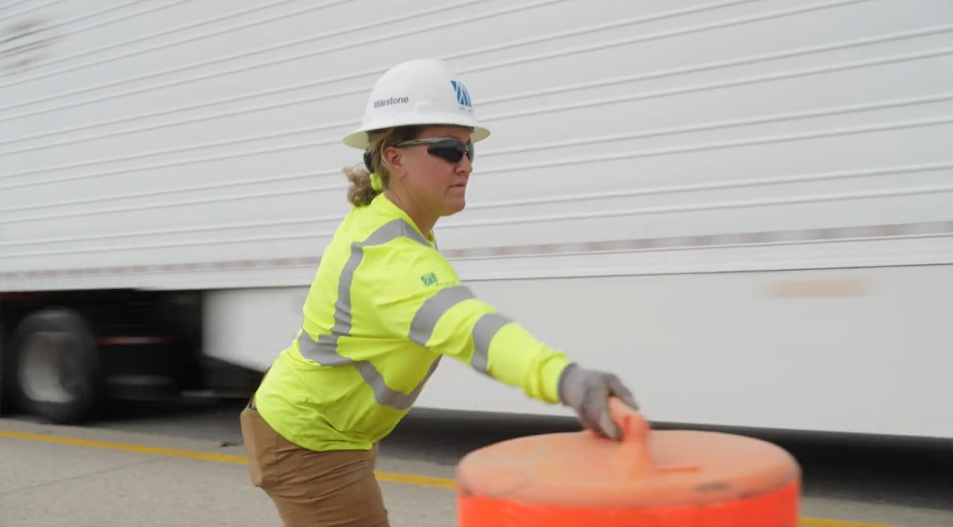 Woman construction worker moving safety barrell on the interstate with semi truck speeding by.