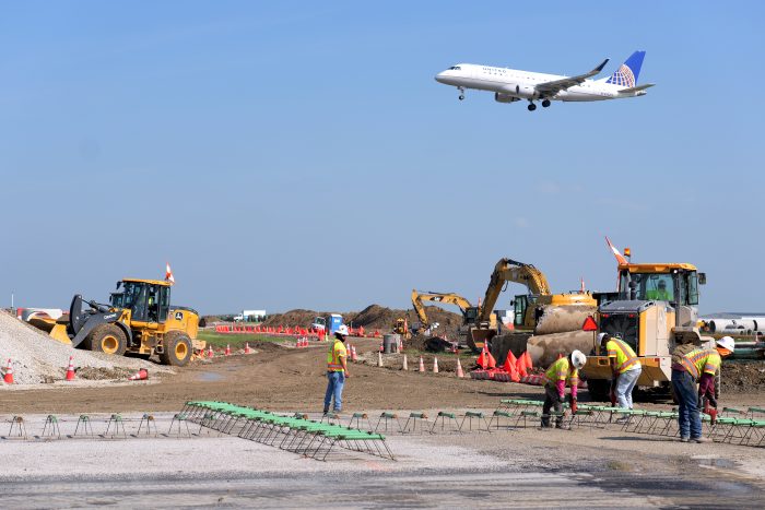 An airplane takes off in the background while Milestone crews construct a new runway.